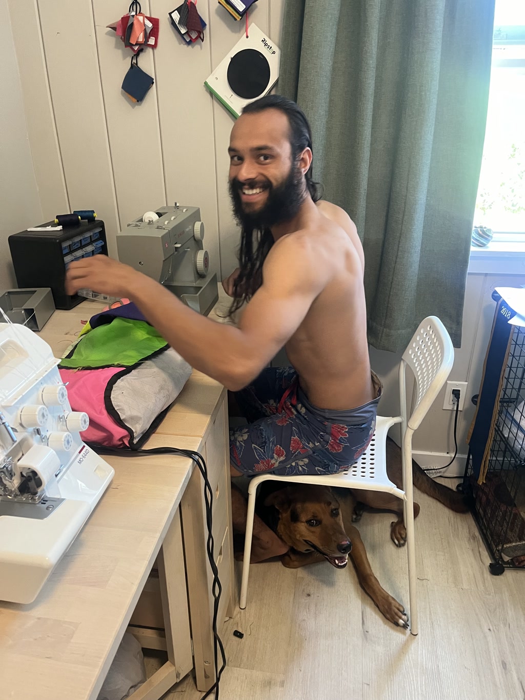 A person with long hair tied back into a ponytail is seated in front of a sewing machine, working on a project. They are not wearing a shirt and are dressed in floral board shorts. Under the chair, a brown dog with a black muzzle appears to be lying down, looking forward with its tongue out as if overseeing the work.