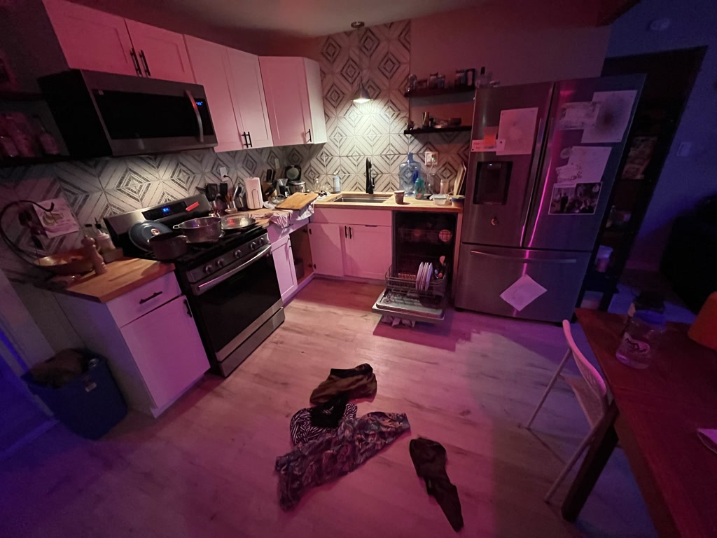 The kitchen displays a vibrant atmosphere with ambient lighting casting a soft glow on the modern appliances, including a stainless steel refrigerator adorned with papers, a stove, and a microwave. The counters are cluttered with various items such as a pan, a pot, cooking utensils, and a chopping board, suggesting a recent meal preparation. The geometric-patterned backsplash adds depth to the space. On the wooden floor, a striped dress and other articles of clothing are strewn about, hinting at an unusual scenario where the cooks may have left their attire behind. Amidst the disarray, there's an intriguing sense of liveliness and untold stories.