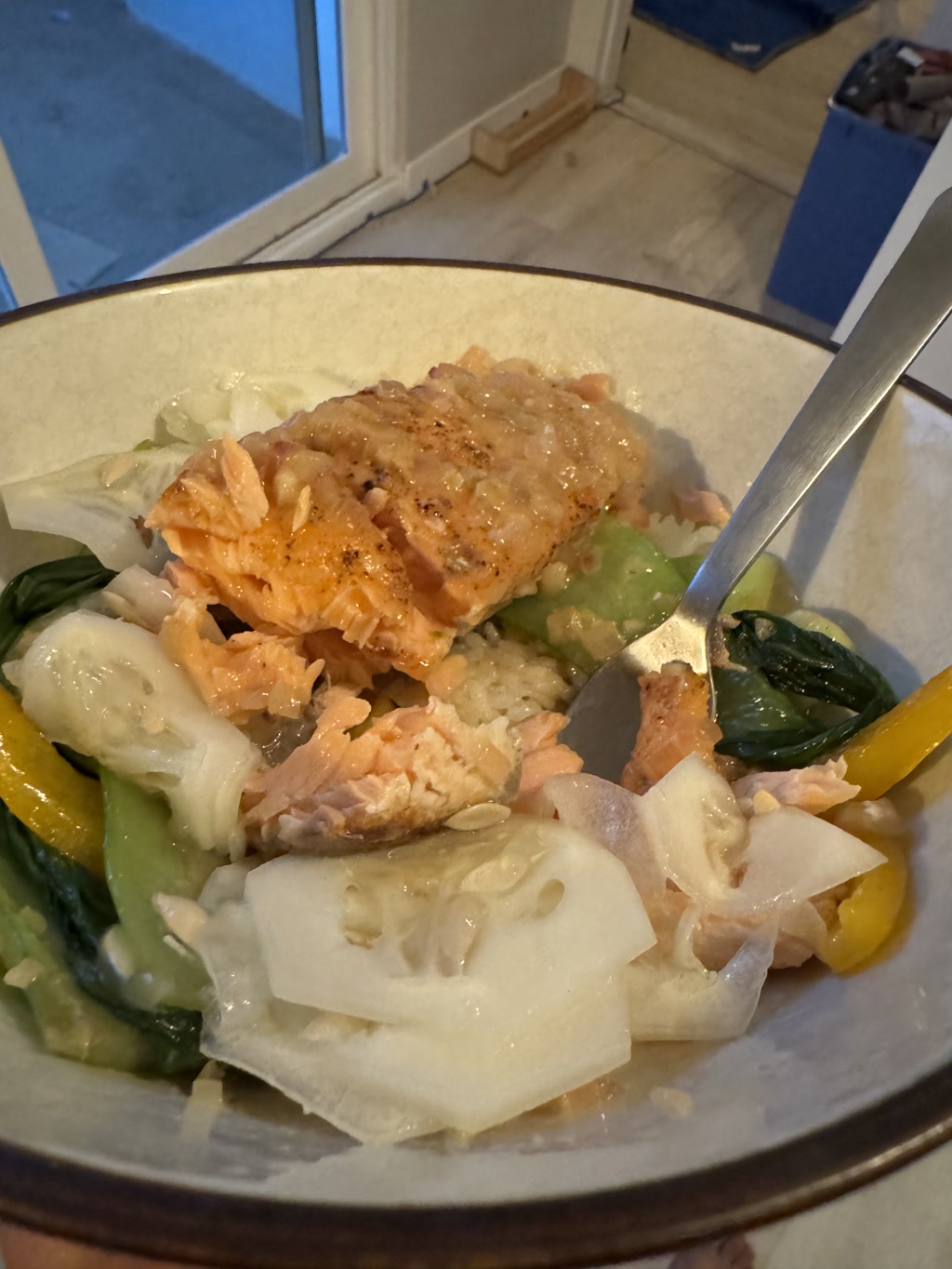 A plate holds a meal comprising a lightly seasoned salmon fillet with a pale golden-brown glaze on top. Below the salmon, a glossy sauce is pooled among a mixture of vegetables including green bok choy, creamy white slices of onion, and bright yellow strips of bell pepper. A portion of the dish shows white rice, and the salmon fillet appears tender and flaky. The background suggests an indoor setting with a view through a door or window.