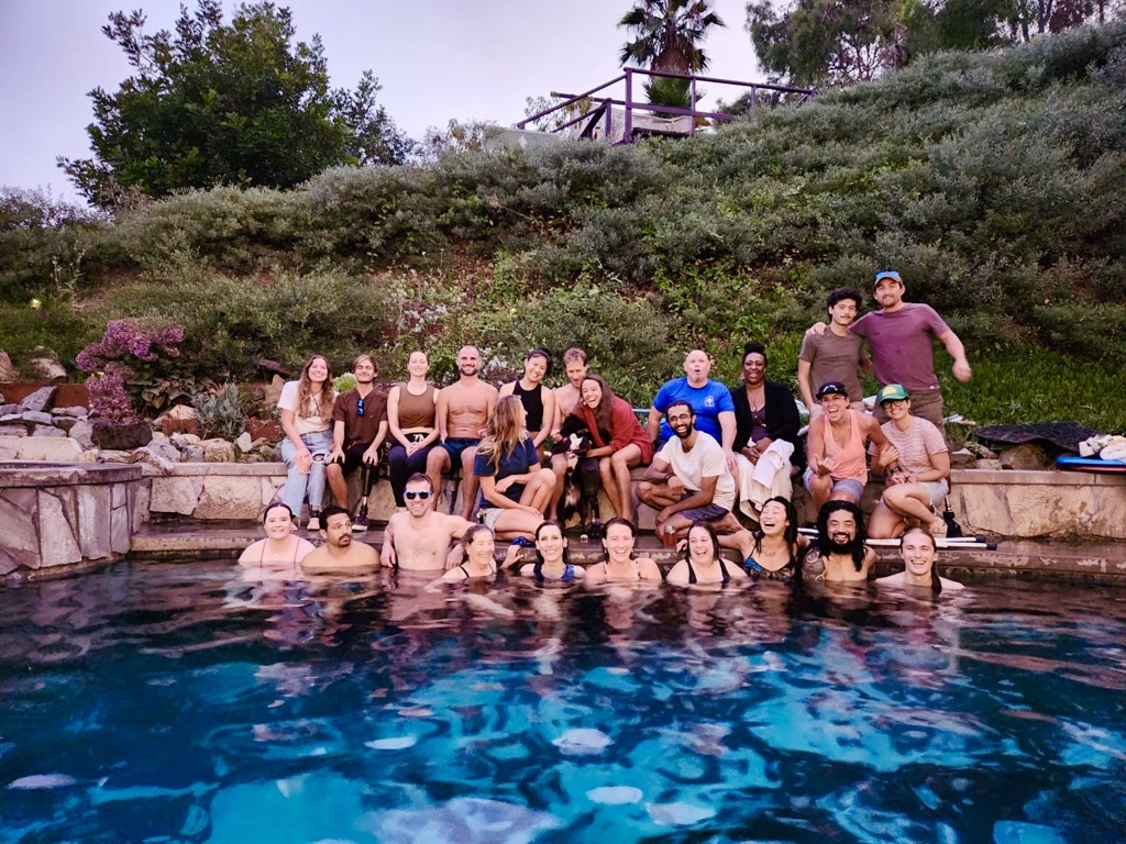 A diverse group of people are enjoying a gathering at a pool. The setting appears to be an outdoor pool area surrounded by natural greenery with bushes and trees. Some individuals are seated on the edge of the pool while others are standing on the poolside. Many in the group are in swimming attire, suggesting a warm, relaxed environment. The group is a mix of genders and ethnicities, and everyone is smiling or laughing, contributing to a joyful atmosphere of camaraderie and leisure.