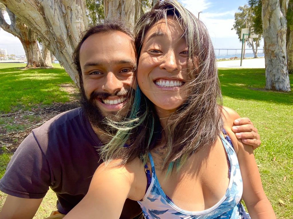 A joyous couple is taking a selfie outdoors on a sunny day. They are smiling broadly, sharing a close embrace, surrounded by green grass and trees. The woman to the right has long hair with a tint of blue at the ends and is wearing a blue patterned tank top. The man on the left has a beard and is wearing a dark T-shirt. Behind them, an expansive park with several trees and a glimpse of a basketball court can be seen, hinting they are at the Embarcadero Marina Park.