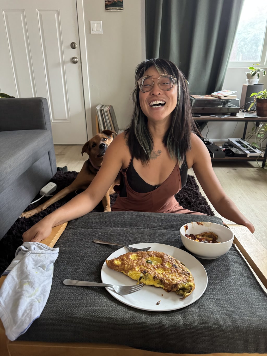 A person with shoulder-length hair and glasses is sitting at a dining table, smiling broadly with their eyes closed. They are wearing a tank top and have several tattoos on their arms. On the table, there's a plate with a large omelette sprinkled with green herbs and a bowl with a reddish-brown fruit compote. A grey sock lies beside the plate. In the background, a brown dog is sitting on the floor, gazing at the scene. The room has a homey feel, with a record player and speakers on a shelf, and a green curtain partially covering a window that lets in daylight.