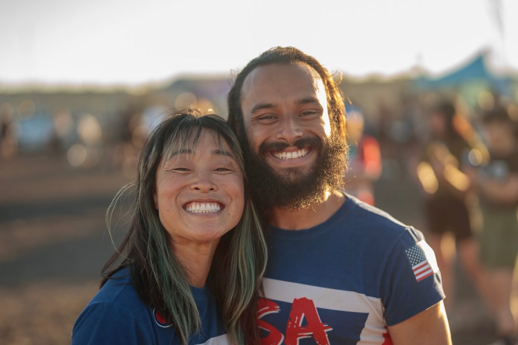 A joyful man and woman are closely posing for a photograph, both offering broad smiles. The woman has long, dark hair with hints of green and is wearing a blue t-shirt with a red collar and what appears to be part of a logo or design. The man has a full beard, long curly hair tied back, and is sporting a blue t-shirt with red and white trim and 'USA' across the front, suggesting patriotic themes likely connected to an American event or celebration. They are both sun-kissed which, along with a blurry crowd in the background and warm lighting, indicates the photo was taken outdoors, possibly at an event like Tracklocross 2024.