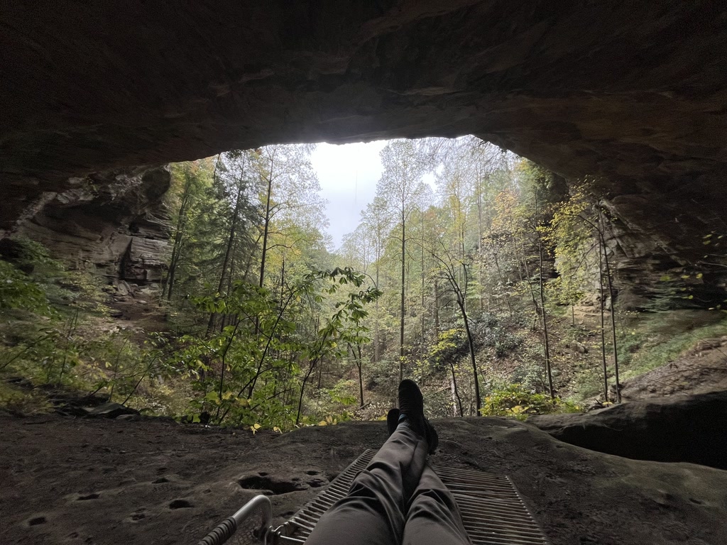 A person is reclining with their feet stretched out, resting on a sandy ground of a cave-like structure that opens up to a lush forest. The view forms a natural frame created by the rocky overhang which reveals towering trees in various shades of green. Leaves are scattered on the ground indicating a season of early fall or late summer. The tranquil scene seems isolated, offering a sense of peace and solitude away from the bustle of everyday life.