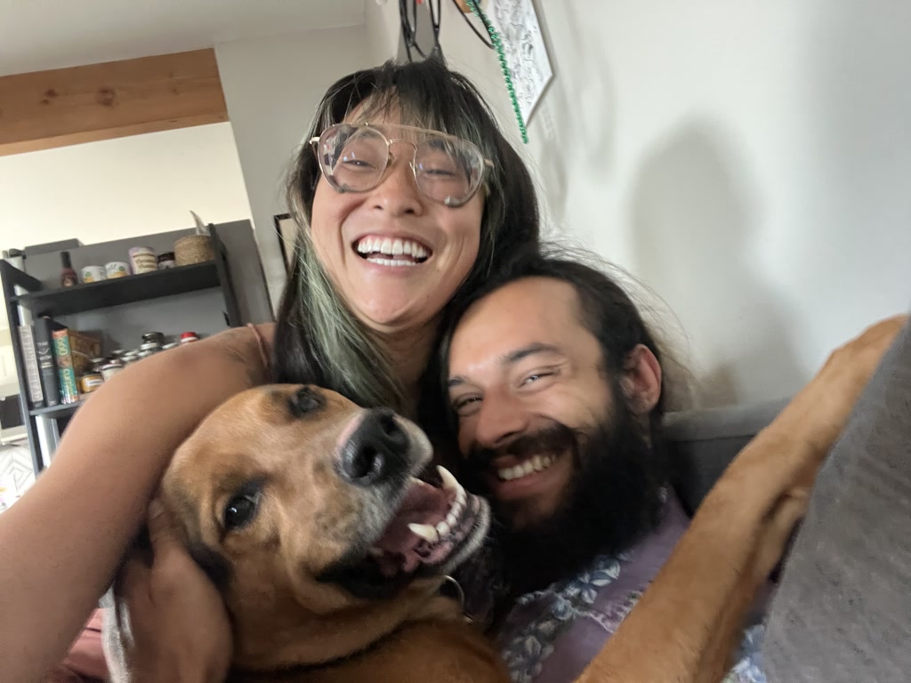 A joy-filled moment with two cheerful adults and a happy dog engaging in a cuddle. The woman is wearing large round glasses and is laughing with her eyes closed while the man has a full beard, long hair, and a broad smile. They both appear to be very happy and content. The dog, caught mid-shake, has its ears flapping and mouth open in what seems like a doggy smile, adding to the overall delightful and affectionate atmosphere of togetherness.