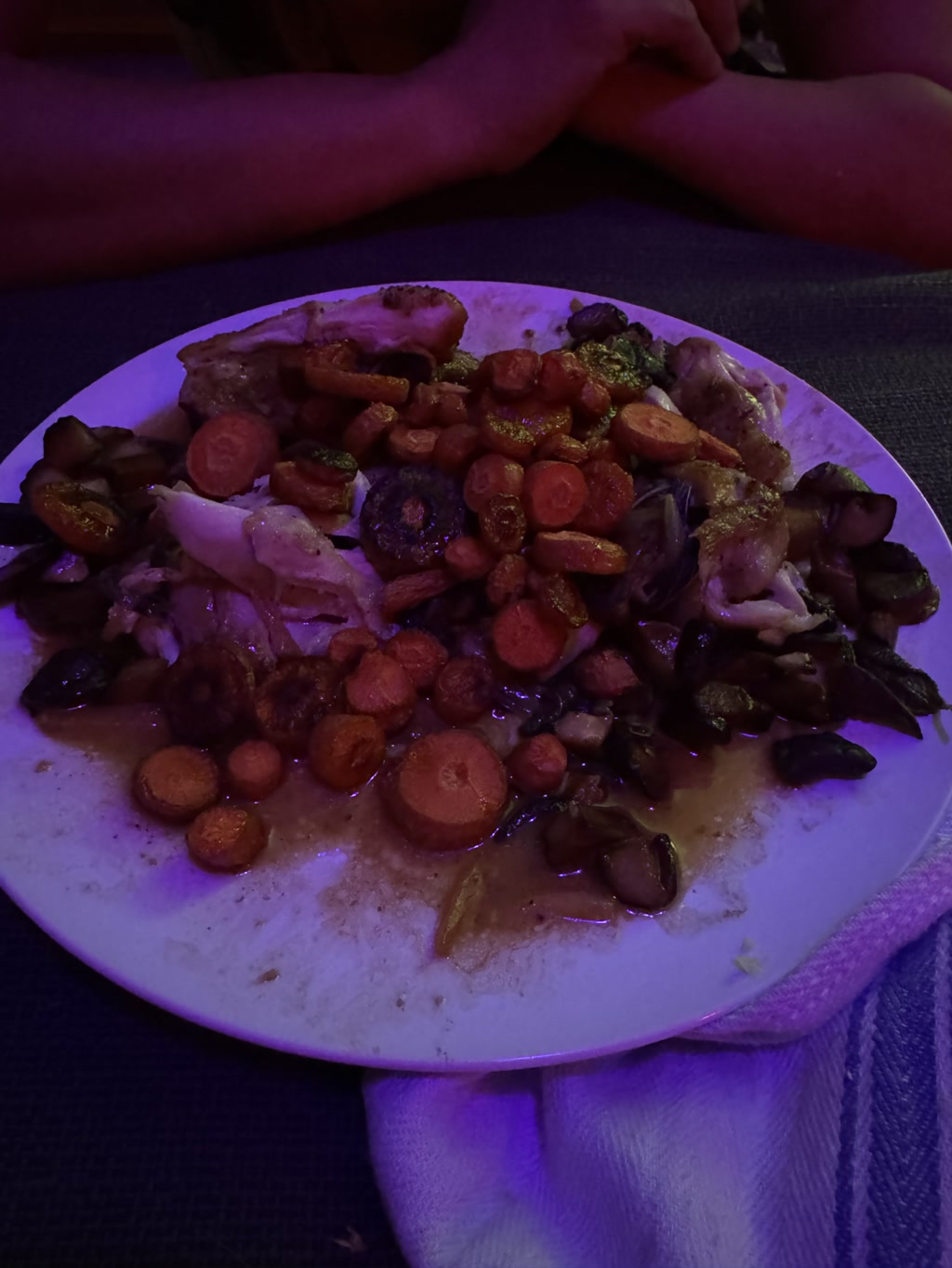 A plate is filled with a mix of roasted vegetables and chicken. The vegetables include carrot slices and pieces of broccoli. The components are coated in what appears to be a savory sauce, suggesting the dish might have been stewed or braised. The lighting is dim with a slight blue and purple hue, possibly indicating the setting is a restaurant with ambient lighting. A person's hands are seen resting on the table, indicating they may be about to eat the meal.