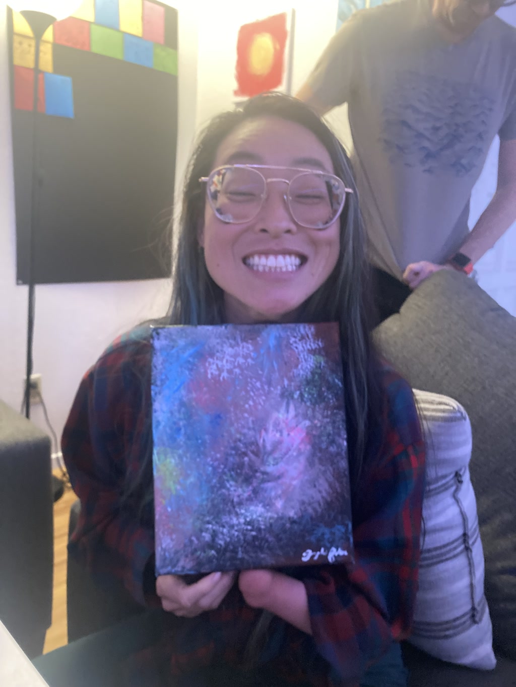 Josie is smiling broadly while holding an abstract acrylic painting. She is wearing clear-rimmed glasses and a plaid shirt, and her long hair is down. The painting features hues of blue and pink with dabbling specks that create an impression of a vibrant cosmic or undersea scene. There's a signature at the bottom of the painting, indicating the artist's mark.