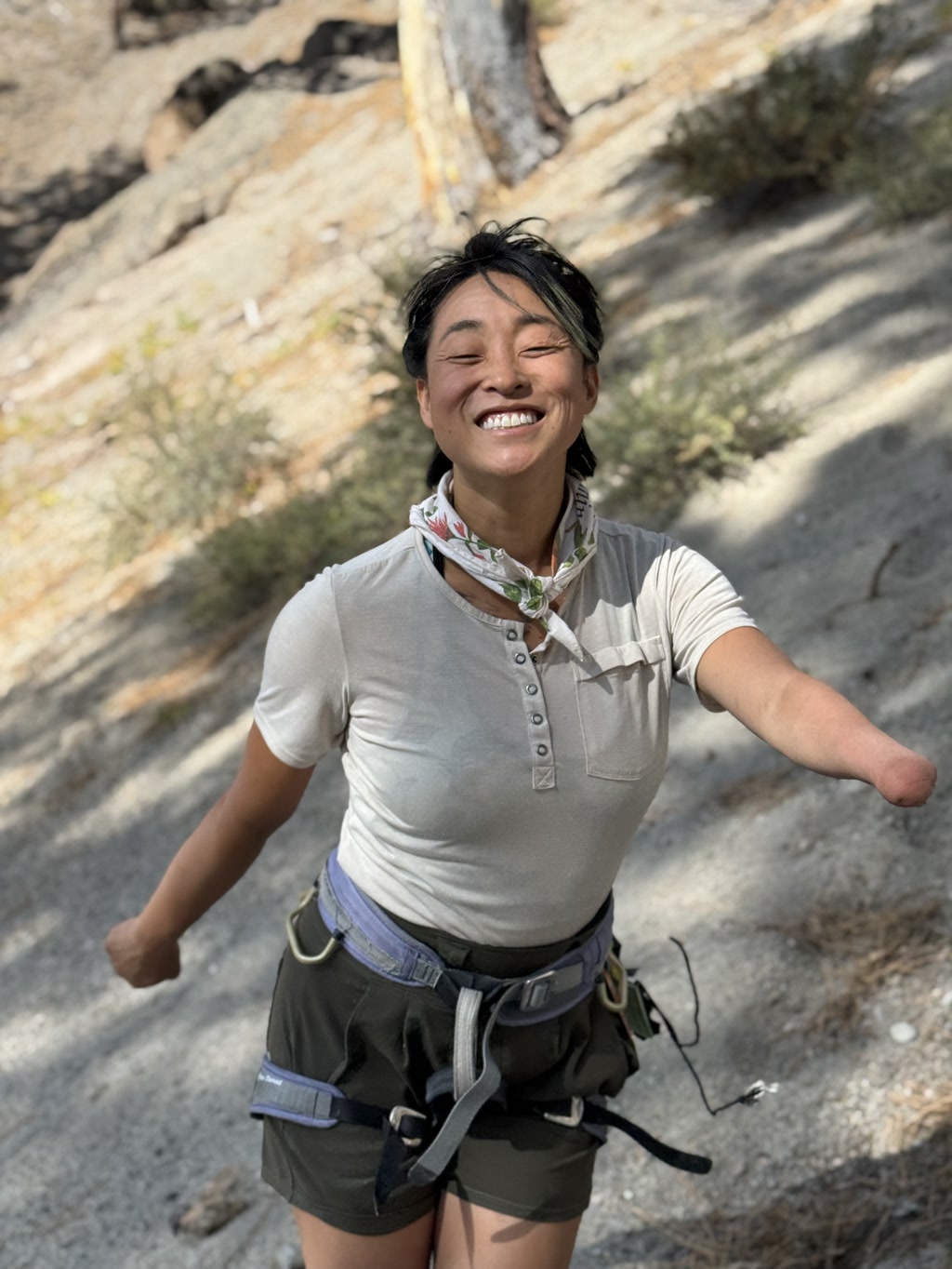 Josie is beaming with excitement in an outdoor setting. She's wearing a casual light-colored shirt with buttons and a bandana tied around her neck. She's geared up with a climbing harness that has multiple loops and carabiners attached to it, indicating she may be preparing for an adventurous activity. Her arms are spread wide, expressing joy or accomplishment, and the background suggests a rocky terrain suitable for climbing. The sunlight casts a glow on her, highlighting her joyful expression.