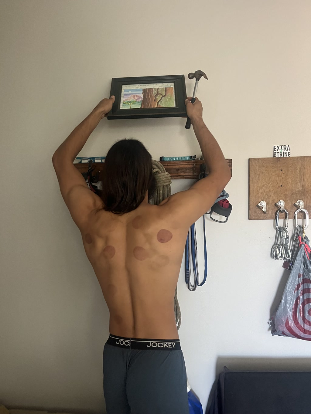 Cj is standing with his back turned, holding up a framed painting of a landscape featuring mountains, trees, and a body of water, using his left hand to hold the frame against the wall and his right hand to position a hammer above a wall-mounted rack filled with climbing gear. On his upper and lower back are round cupping therapy marks. To his right are hanging carabiners labeled 'EXTRA STRING' and a bag draped over a piece of workout equipment. He is wearing dark underwear with the brand visible on the waistband.