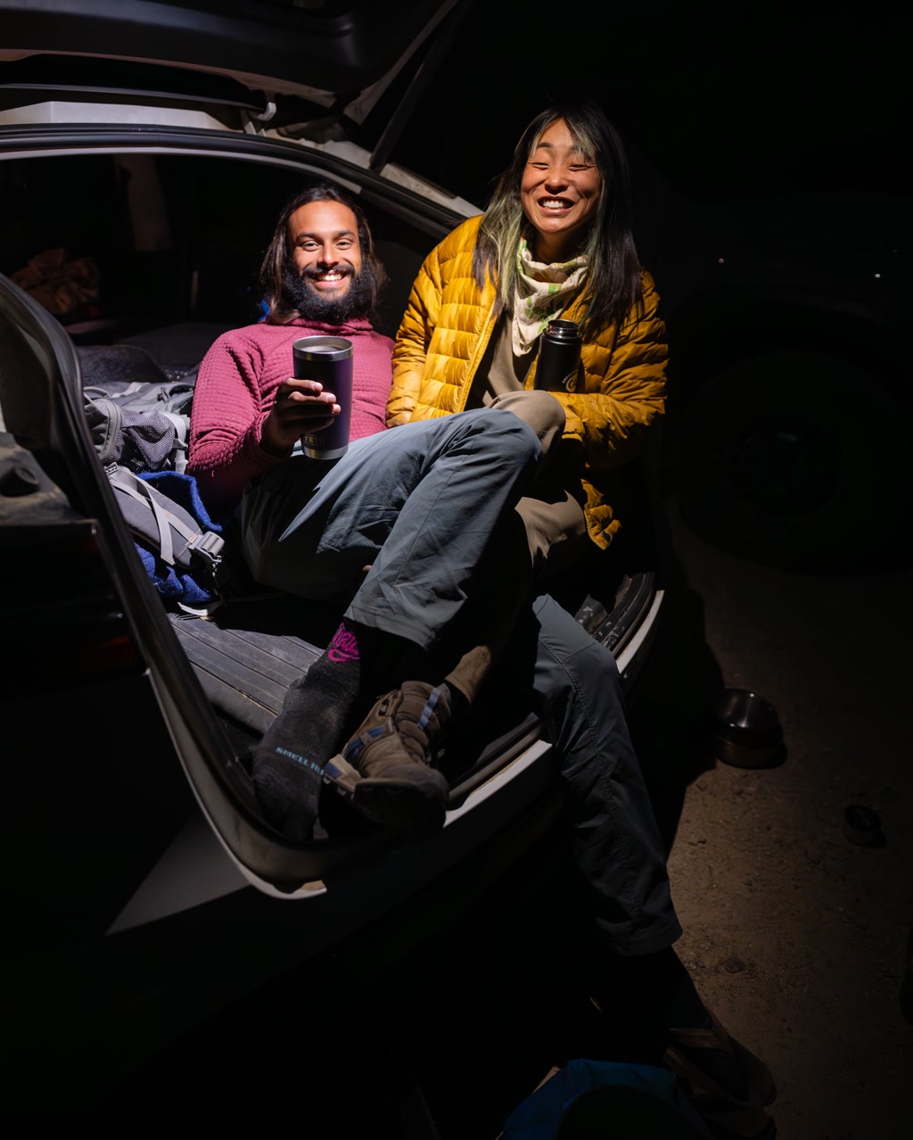 Two individuals are cozily seated in the rear of a vehicle, presumably during a camping trip. The person on the left is sporting a pink sweater and holding a mug, with a contented smile and a relaxed demeanor. The person on the right is wearing a vibrant yellow jacket and also holds a mug, radiating warmth and joy with their smile. Both of them are wearing layered clothing suitable for outdoor activities. They're surrounded by an assortment of camping gear and personal items. Their footwear and the vehicle's interior suggest they are in a wilderness setting, enjoying a break from the day's activities by sipping what appears to be warm broth from their mugs.