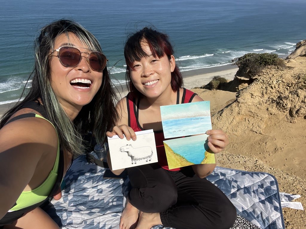 Josie and Dana are sitting on a cliff overlooking the beach. The ocean waves break gently on the shore below them. They are both smiling widely, capturing a joyful moment together. Josie, with her sun-kissed hair and large sunglasses, holds a drawing of an animal that resembles a polar bear, while Dana, sporting a red top, presents a seascape painting divided into two separate pieces which, when aligned, create a continuous panoramic depiction of a beach scene. The brilliant hues of blue and sand in Dana's artwork complement the natural scenery behind them. A patterned blanket is spread beneath them, and the clear sky above indicates a beautiful, sunny day.