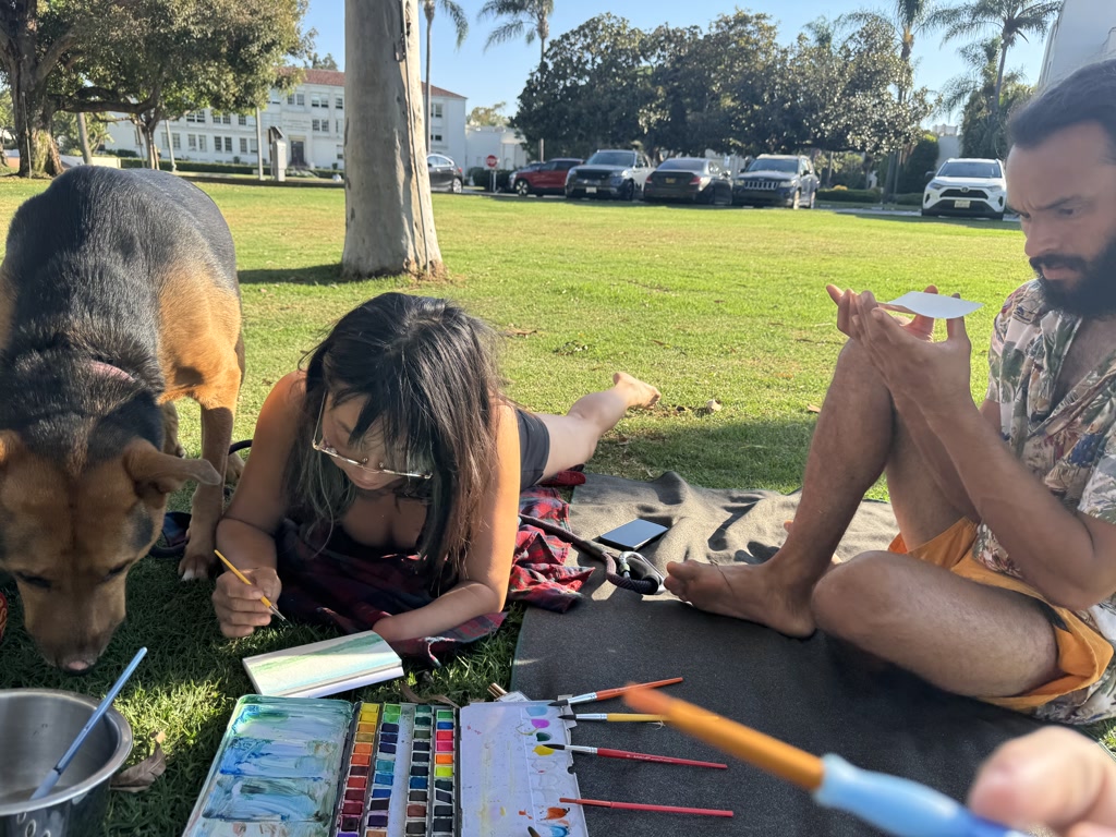 A man and a woman are engaging in outdoor activities on a sunny day in a park with grass and trees. The woman is lying on her stomach on a blanket, concentrating on a notebook with a pen in hand, with painting supplies like a palette and various brushes spread around her. To her right, a man is sitting on the blanket, intensely focusing on a small white piece of paper that he is holding up with both hands. It appears he might be painting on it, with the sun casting shadows over his work. In the foreground, a dog with a brown and black coat is casually sniffing around, looking away from the people. The scene is relaxed and creative, with a peaceful atmosphere.