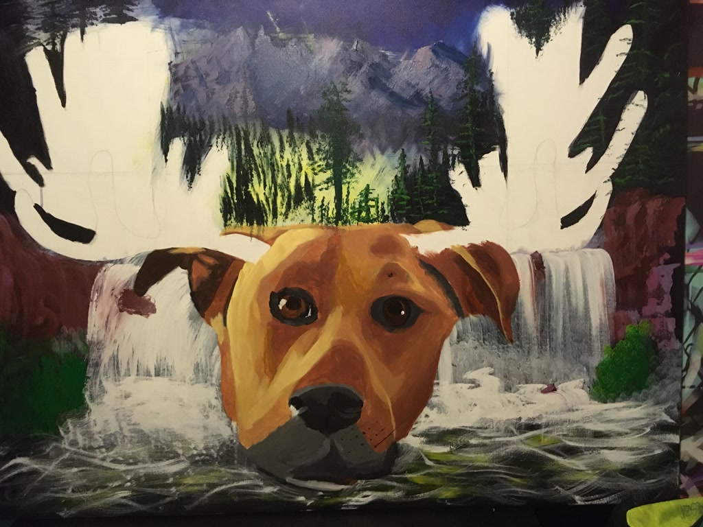 An unfinished acrylic painting features a dog with warm brown and golden fur, with the notable addition of moose antlers outlined but not yet filled in. The dog's large, expressive brown eye and perked ears draw attention. In the background, a picturesque scene unfolds, showcasing mountains shrouded in mist, dense green forests, and a cascading waterfall that seems to flow around the dog's head. The waterfall is depicted with a dynamic sense of movement, as whitewater tumbles over rocky edges into a serene pool below. The colors are vibrant and rich, creating a lively contrast between the earthy tones of the dog and the cool blues and greens of the natural landscape.