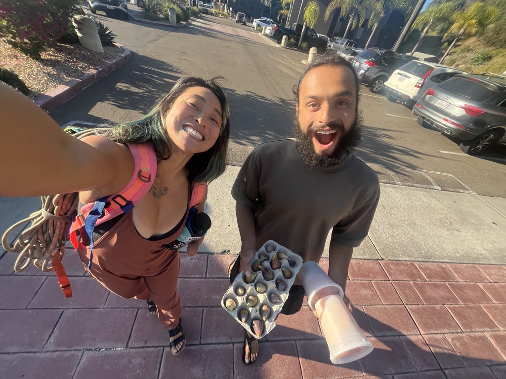 Two joyful people are seen outdoors, likely in a parking lot with cars in the background. The person in the foreground, to the left, appears to be a woman with long hair dyed with a bluish tint, broad smile, and is wearing sporty, outdoor clothing and a pink climbing harness with a rope coiled over one shoulder. She is extending her arm possibly to take the selfie. The individual to the right is a man with a full beard, wide-open mouth as if exclaiming in surprise or excitement, and dressed in a dark brown T-shirt. He is holding a curved tray filled with figs in one hand and a large transparent plastic cup with a pinkish liquid in the other. Both individuals seem to be in high spirits, and the scene conveys a sense of adventure and camaraderie.