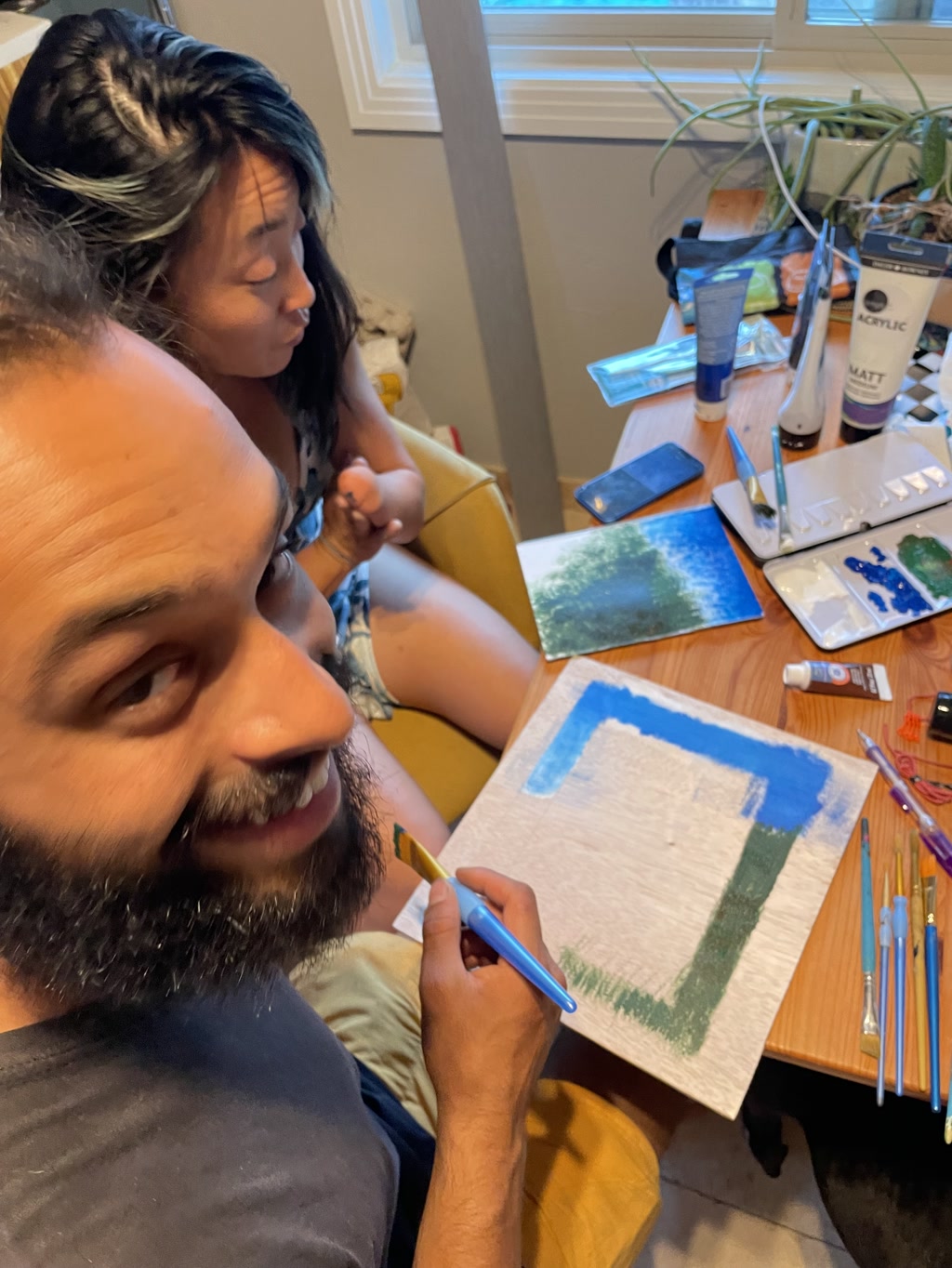 A couple is seated at a table engaged in a painting activity. The man smiles at the camera, holding a paintbrush, with a partially painted canvas in front of him showcasing a bold blue sky above a green area that suggests the top edge of a forest. The woman, focused on her work, is adding detail to a smaller canvas. Her canvas features a lush green representation of a forest and seems as if it could seamlessly fit beneath the blue and green section of the man's canvas, completing the outdoor scene. Paints, brushes, and other art supplies are scattered on the table, evidencing the creative process they are both involved in.