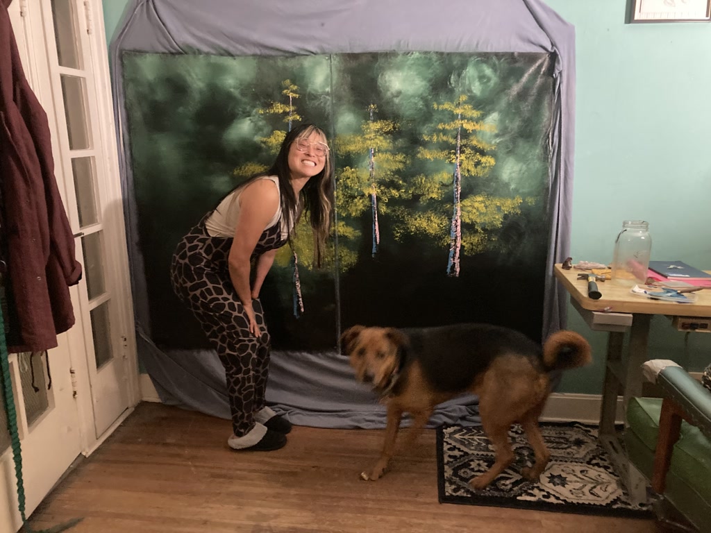 Josie poses with a playful stance to her left, flashing a bright smile while wearing an animal print jumpsuit. Behind her is a large life-size canvas featuring the beginnings of an acrylic forest scene, boasting a variety of green hues and hints of yellow that suggest sunlight filtering through the trees. To her right, Moose, a medium-sized, furry dog with a black and tan coat, appears in motion, possibly walking past or turning away, with its tail mid-swing. The room has wooden flooring and pale teal walls, with a table holding various items to the right, including a jar and some papers.