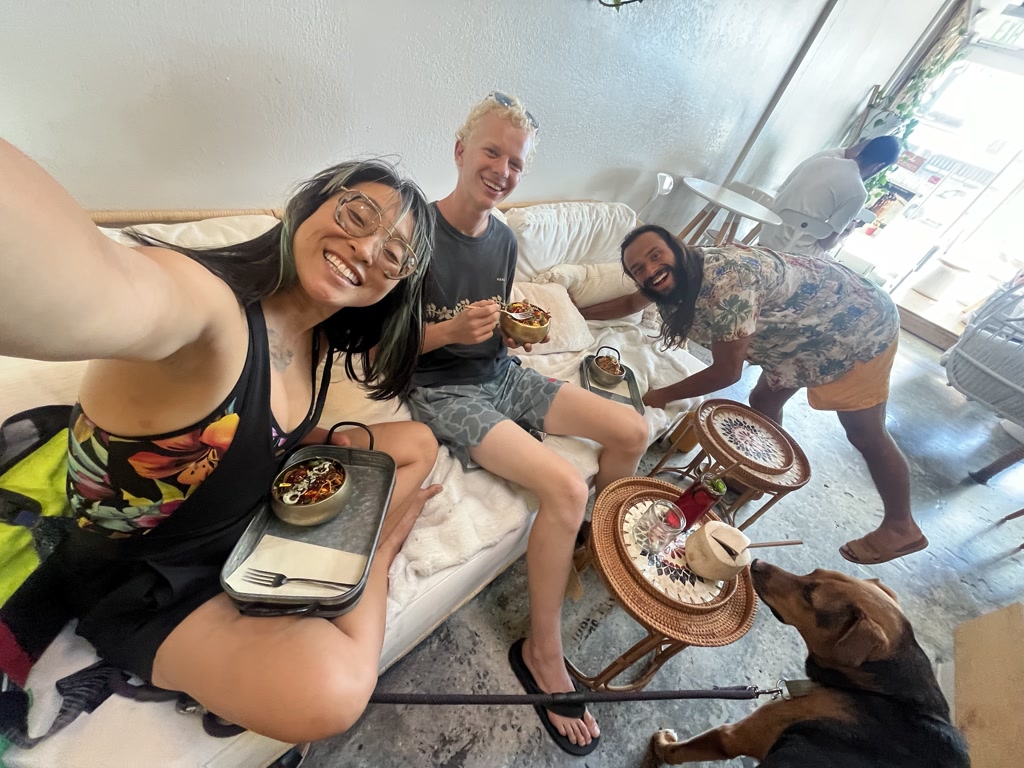 Three friends are gathered in a cozy indoor setting, enjoying a casual meal together. Josie, on the left, is smiling brightly at the camera while holding a tray with a bowl of food; she wears glasses and a sleeveless top with a colorful print. Ollie, in the middle, has a cheerful expression and light-colored hair, casually dressed and also holding a bowl of food. CJ stands on the right, leaning towards the camera with a playful smile and wearing a floral-patterned shirt and shorts. All of them are seated or kneeling around a low table adorned with a small plant, and there is a dog, possibly named Ollie, who is engaged with one of the friends, its attention directed towards a coconut-like object on the table. The environment has a laid-back vibe, with a white couch and natural light streaming in from a door in the background, suggesting a relaxed and friendly mealtime encounter at a place they refer to as Sustainabowl.