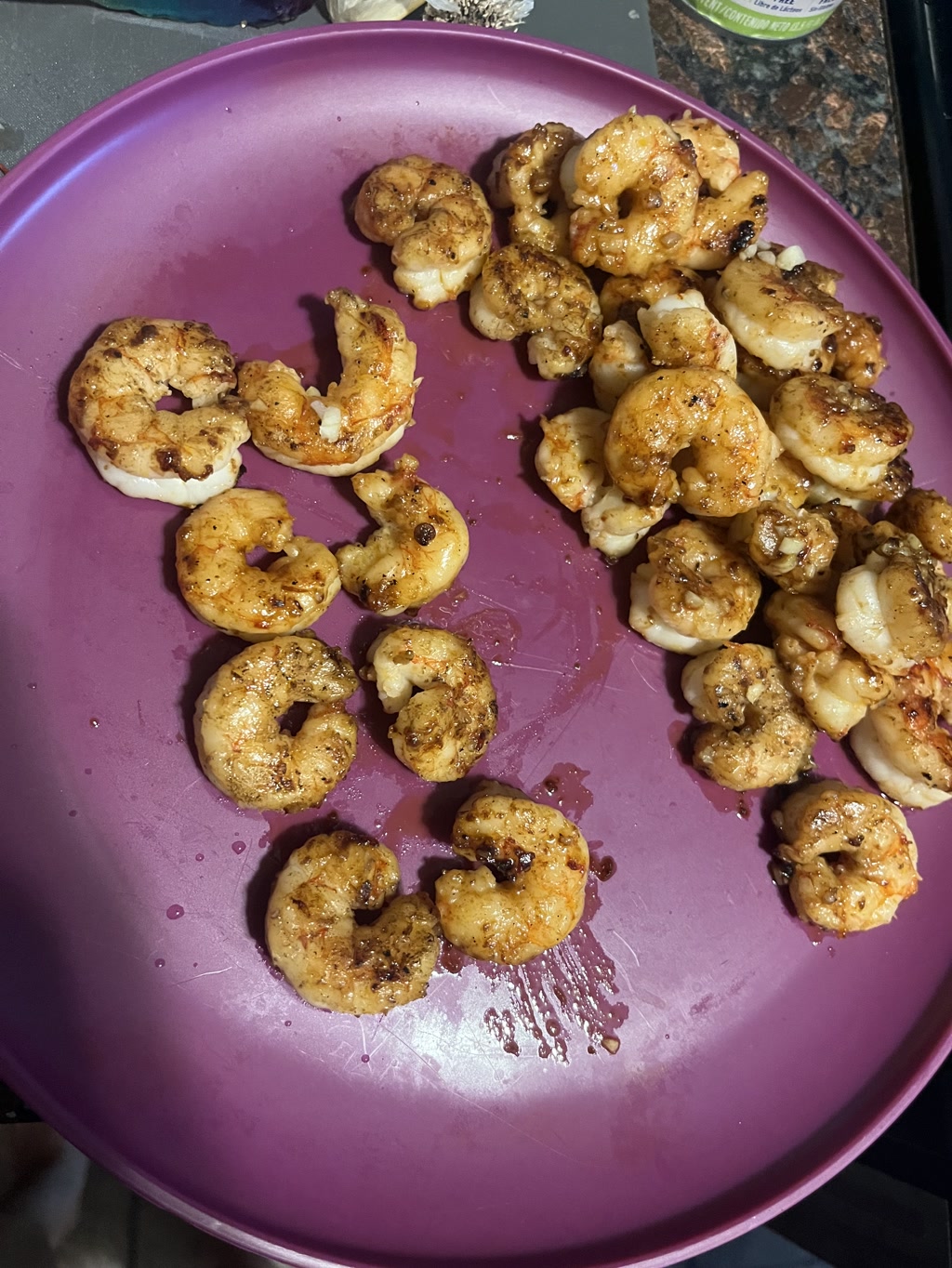 Butter-fried shrimp are neatly arranged on a purple plate. On the left side of the plate, shrimp are meticulously lined up to form the letters C and J, representing initials or a special meaning. The shrimp forming the C are curved into a crescent, whereas the ones forming the J have a top curve and a longer tail. To the right, another pile of shrimp is gathered, cooked to a golden color indicative of being fried in butter, with slight char suggesting a high-temperature searing.