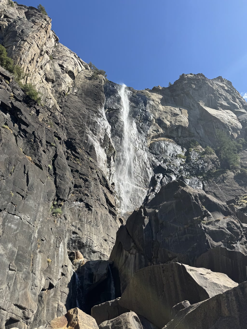 A majestic view of Bridalveil Falls, featuring cascading water flowing gracefully down a rugged cliff face. The waterfall is surrounded by large, steep rocky outcrops with sparse vegetation clinging to the crevices. Sunlight dances on the water and the granite surfaces, highlighting the natural textures and creating a play of light and shadow.