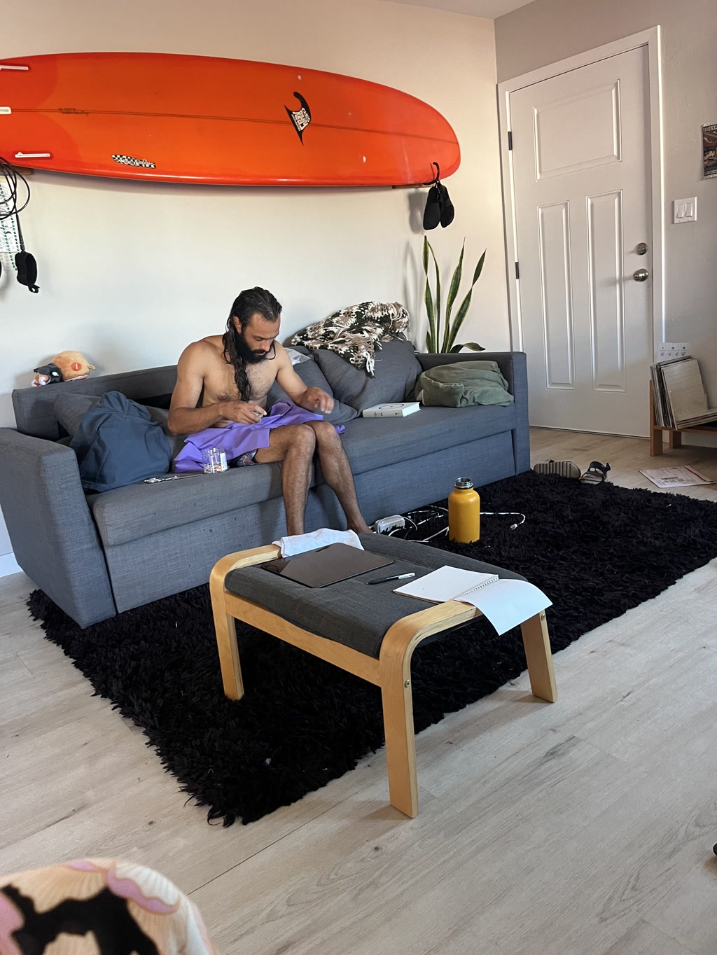 A person is sitting on a gray couch, focused intently on sewing a purple piece of fabric, which is likely part of a hoodie. The setting appears to be a cozy living room, and the person has a long beard and hair tied back. Behind them, on the wall, hangs an orange surfboard with black fins and a white shark logo. A speckled dog is resting on the couch, partly covered by a blanket. A small wooden bench with a cushion is in front of the couch, featuring a couple of papers that appear to have writing on them. The room also includes a door to the right, a plant, and various other items that suggest the individual enjoys a relaxed and active lifestyle.