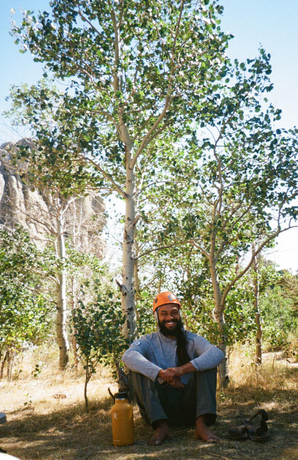 A person with a noticeable smile is seated on the ground, surrounded by trees with green leaves. The individual has a full beard and long hair, is barefoot, and is wearing a gray long-sleeve shirt, blue pants, and an orange helmet. Next to them is a metal water bottle with a visible logo, and a pair of sandals rests on the ground nearby. The environment suggests an outdoor, possibly adventurous setting with clear skies and hints of rock formations in the background.