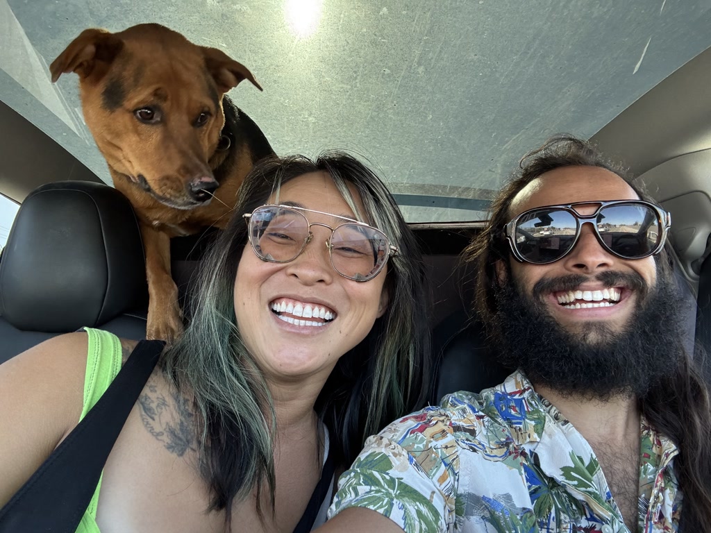 A joyous moment is captured with two individuals and a dog in the interior of a vehicle. The person on the left is wearing large, clear-framed glasses and has a grin with teeth prominently displayed, showing happiness. Featuring dyed hair with shades of green and a visible tattoo on the arm, this person is dressed in a lime green sleeve and a patterned garment. The individual on the right exhibits a full beard, with hair past shoulder length, and is wearing large, reflective aviator sunglasses. A tropical patterned shirt adds to the relaxed, joyful vibe of the snapshot. Between them, a medium-sized dog with a brown coat and curious expression is perching over the back seat, its attention seemingly directed outside the vehicle or towards the front. All appear content and oblivious to the dog's urgency.