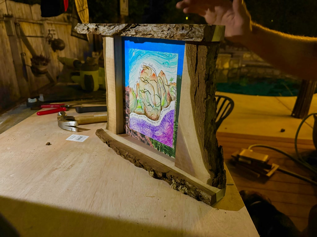 A handmade gift composed of a unique wooden frame with natural, raw edges that display visually different colored layers in the wood grain. Within the frame lies an artistic acrylic background illustrating an overview of a lush forest. Superimposed on this background is a watercolor painting that depicts a mystical, rocky crag teeming with green moss. A heartwarming quote is woven into the artwork, offering a reminder of unrecognized beauty in familiarity.