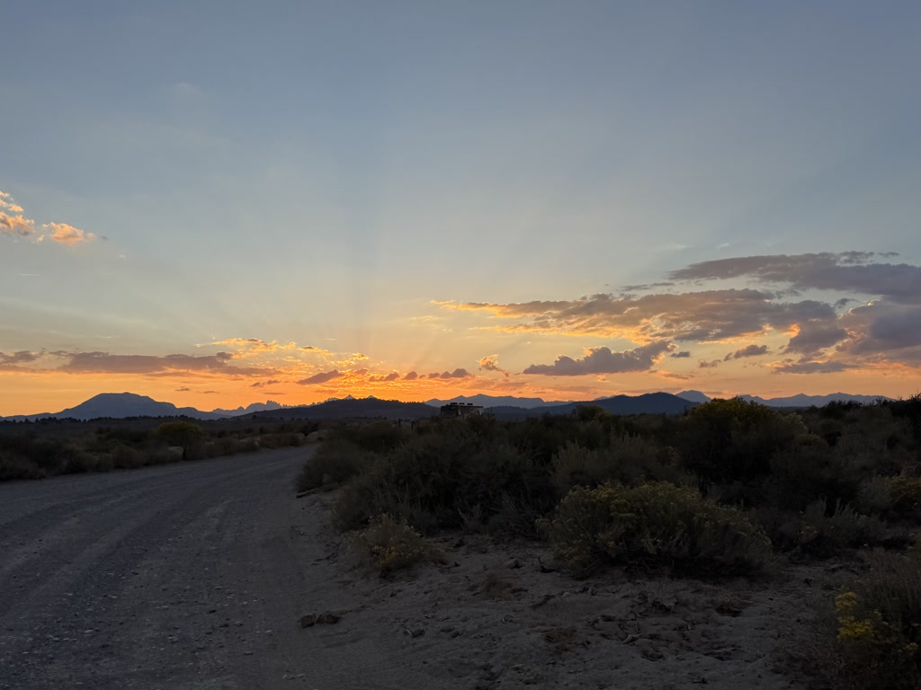 The sun is setting behind a mountain range, casting warm hues of orange and yellow across the sky which are contrasted by soft shades of blue and gray. Below the sky, a rough dirt road meanders through the scene, flanked by scrubby desert vegetation and small plants which are common in arid landscapes. The scene is devoid of any human presence, evoking a sense of serene isolation as the day comes to an end.