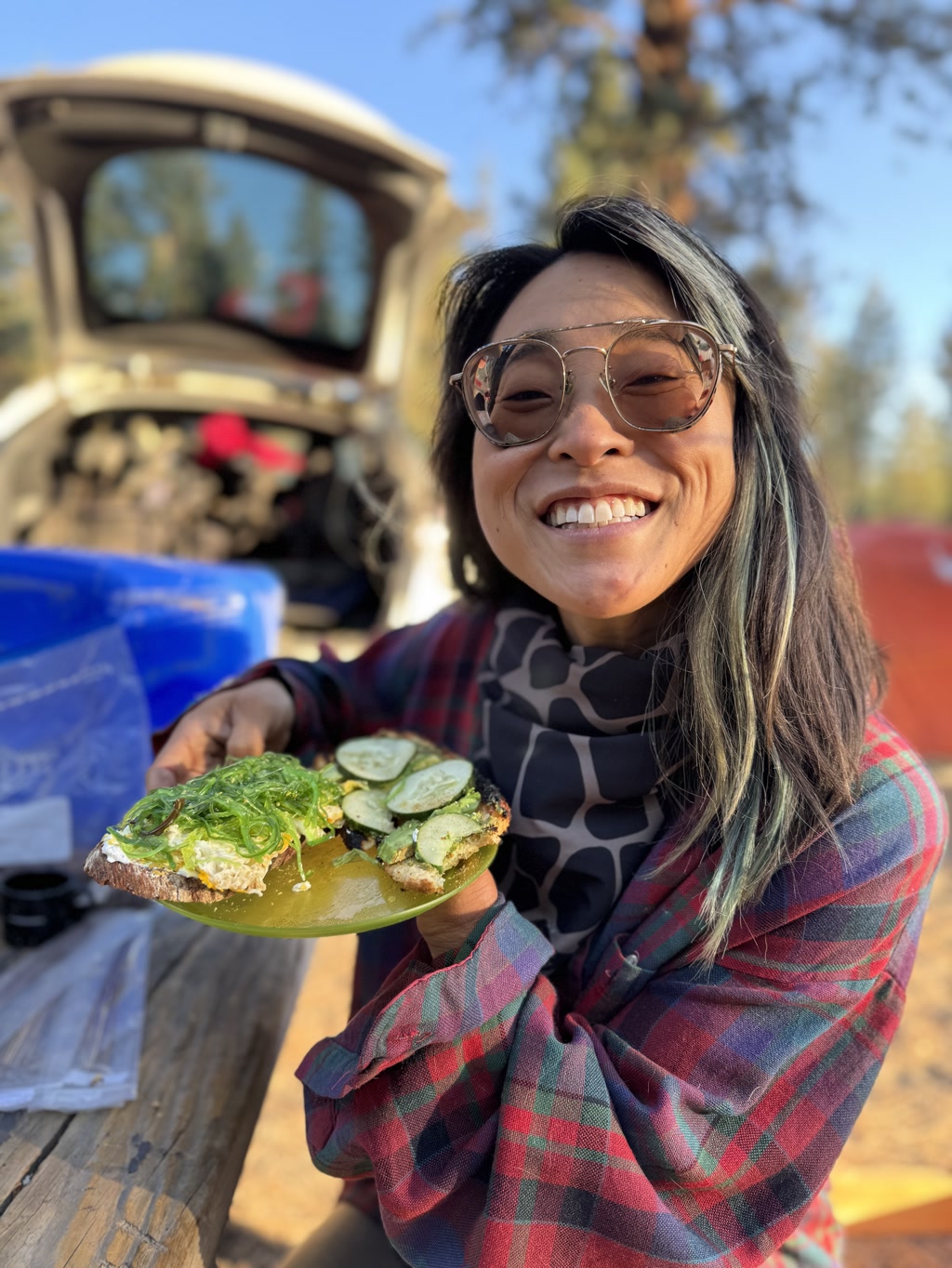 Josie is smiling broadly and holding out a plate with a healthy and appetizing meal. She's wearing round-framed glasses and her hair has streaks of blue. She is dressed in a flannel shirt with a plaid pattern of red, blue, and green, layered over a top with a darker abstract design. Behind her, one can see a sunny outdoor setting with pine trees and the blurred silhouette of a vehicle. The plate contains a slice of fresh bread topped with a generous layer of avocado, a seaweed salad, cucumber slices, and what appears to be eggs. The breakfast looks freshly prepared and suitable for a camping adventure.