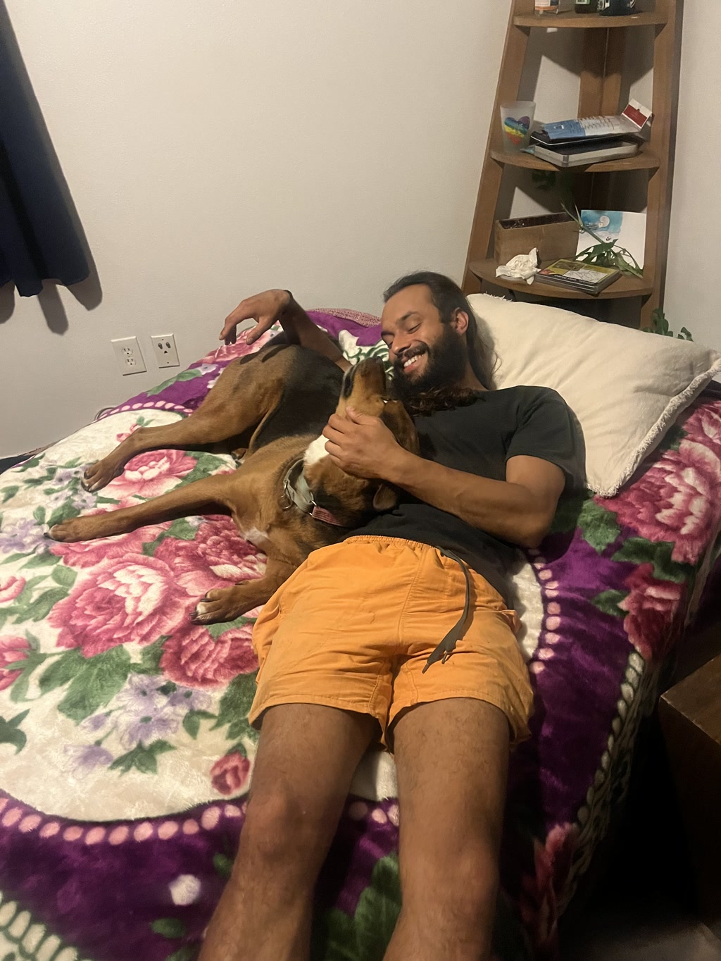 A man with a beard and long hair is lying on a bed covered with a floral-patterned blanket. He is wearing a dark t-shirt and orange shorts. The man is smiling and looking at a large brown dog lying beside him. The dog, seemingly a mix with a glossy coat and a white patch on its chest, is turned towards the man, and they appear to be gazing into each other's eyes in a moment of affectionate connection. There is a bookshelf in the background with various small items and books on it.