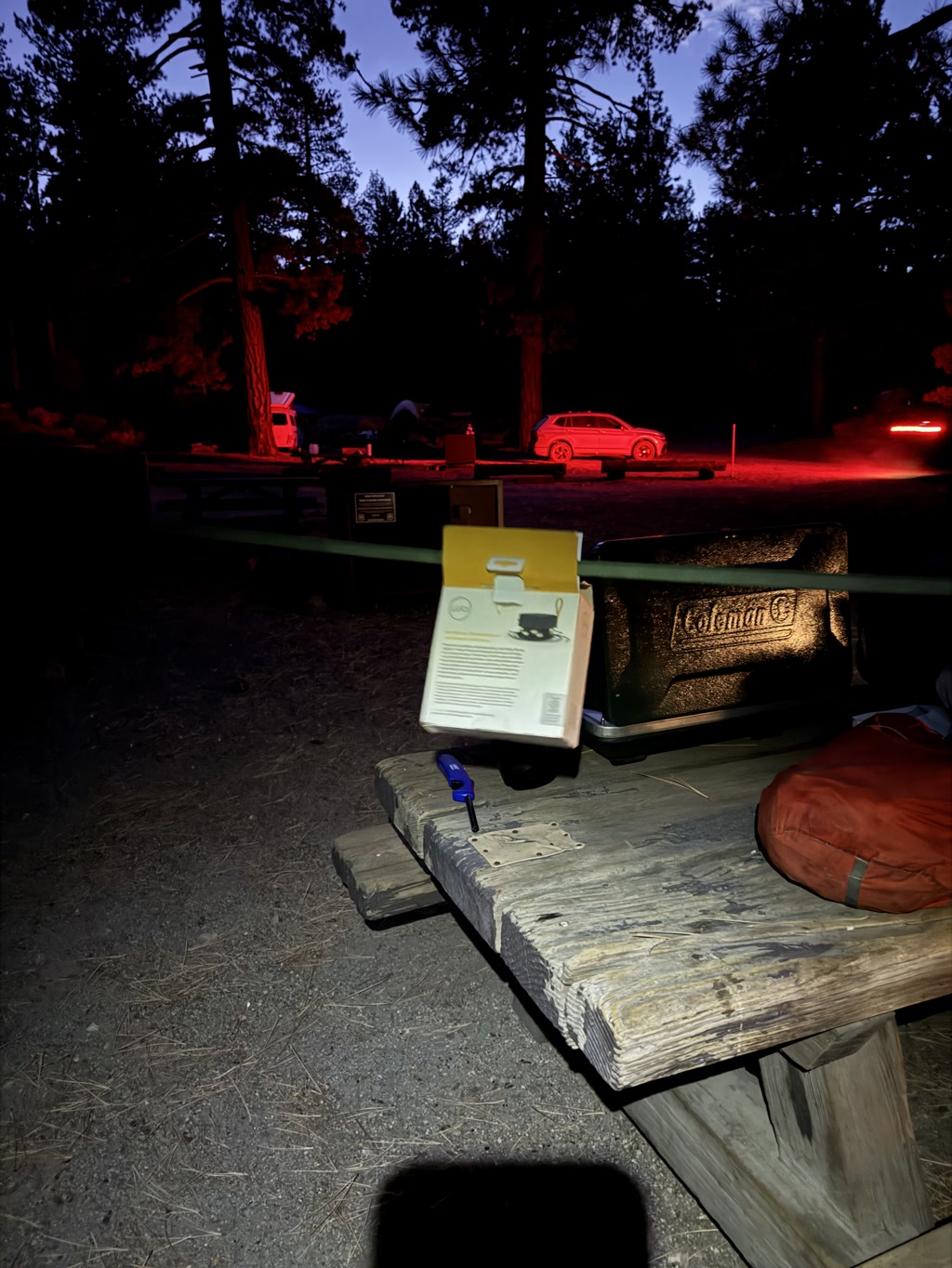 It's a nighttime scene at a campsite with tall trees surrounding the area. A carton is resting on the edge of a wooden picnic table, with the bottom portion lifted off the table's surface. A blue object, possibly a lighter, is on the table near the carton. In the background, a camp grill, a red sedan, and a car with its brake lights on are visible. The ambient light is faint, but the brake lights cast a red glow. The carton on the table appears to have text and imagery on its side.