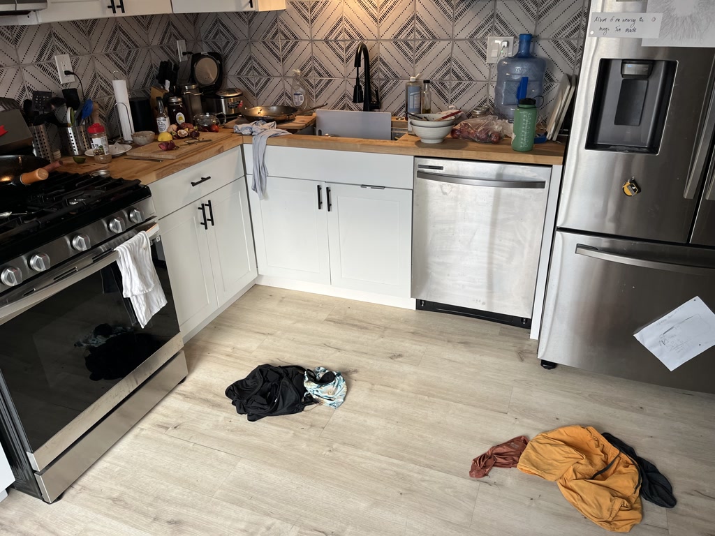 A kitchen with white cabinetry and a patterned backsplash. The countertops are cluttered with various cooking utensils, jars, a toaster, and a cutting board with an onion and sweet potato on it. A cloth hangs from the oven door handle. The dishwasher is ajar, and a stainless steel refrigerator is equipped with a water and ice dispenser. A handwritten note is attached to the fridge. On the wooden floor lie several piles of clothes.