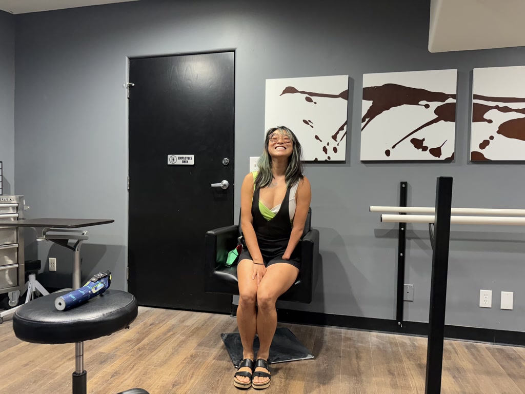 A smiling person, Josie, is seated on a black chair in a room with gray walls. Behind her are three square white canvases adorned with brown abstract patterns on the wall. To her side is a small medical equipment table with metallic tools, and in front of her is a black stool with a blue cylindrical object resting on it. The door beside her has a sign that reads 'EMPLOYEES ONLY'. The overall setting suggests she could be in a medical office or a prosthetic fitting room.