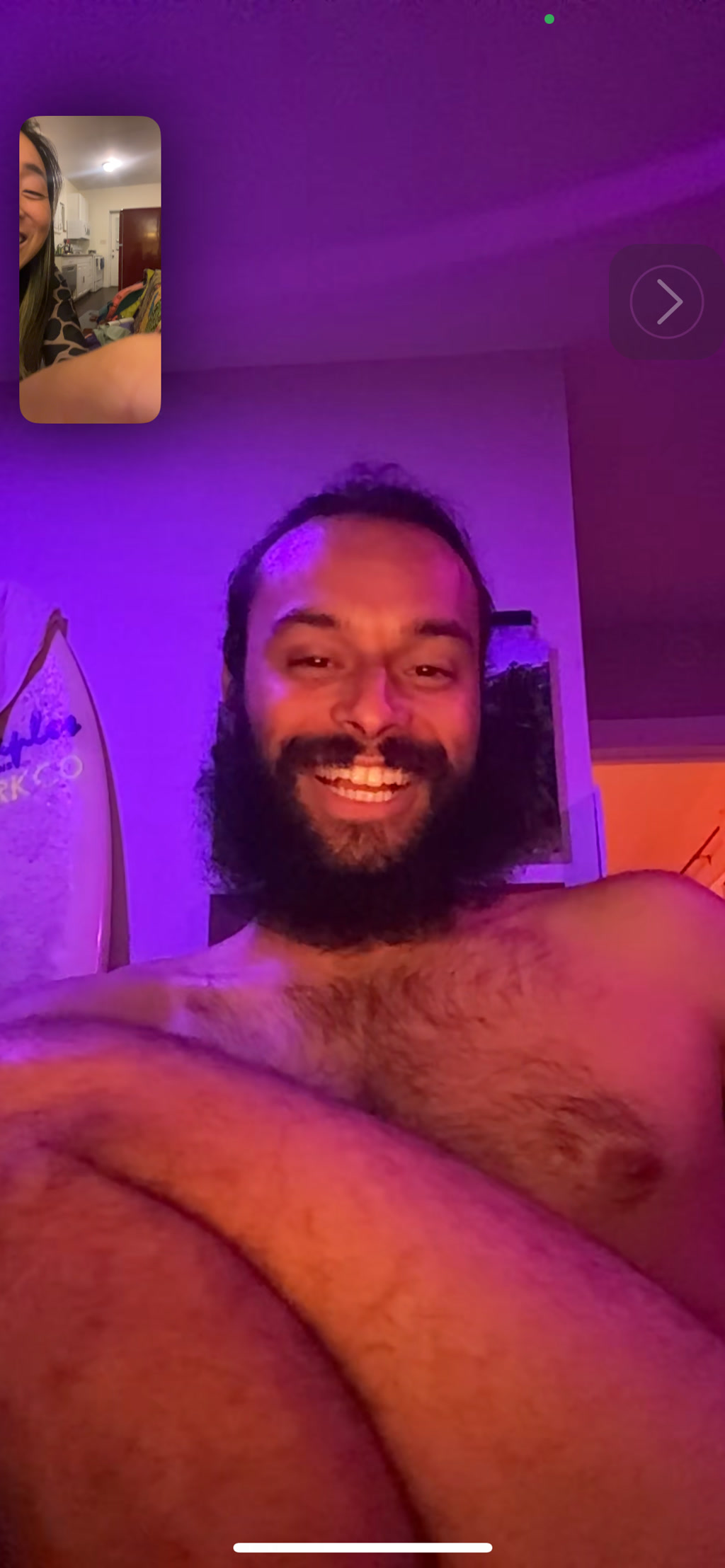 A screenshot from a FaceTime call depicting a person with a beaming smile and long hair. The individual is visible from the mid-chest upwards and their bare shoulders are exposed, creating the illusion that they might not be wearing clothing, although this isn't confirmed within the frame. The scene is bathed in purple and orange lighting, adding to the casual and comfortable atmosphere of the interaction. There's a mirror in the background reflecting a dimly lit room. A small inset window shows another person engaged in the call, their expression one of laughter and enjoyment.