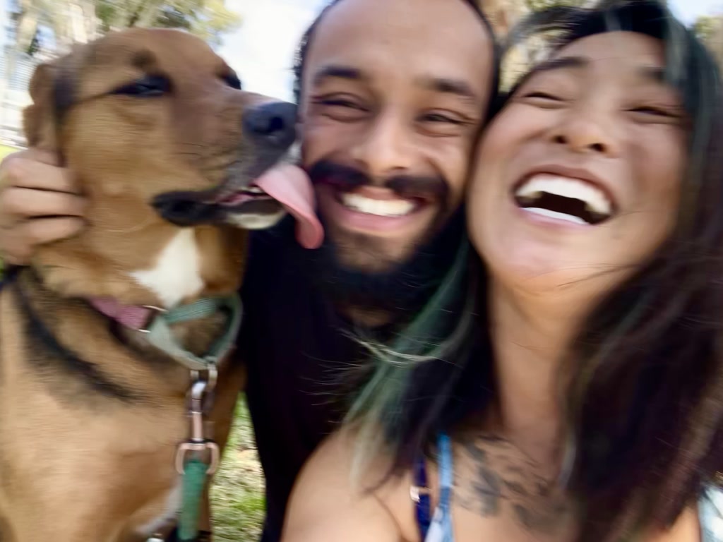 A scene filled with joy as a dog with a brown coat and a lolling tongue licks the face of a smiling man with a beard. The man's face is snuggled closely to a woman's cheek, and both are laughing heartily. Their expressions exude warmth and affection. The dog wears a collar, hinting that it is a beloved pet. All facial features, while distinguishable, are somewhat out of focus due to the motion present in the moment, contributing to the candid and dynamic feel of the scene.