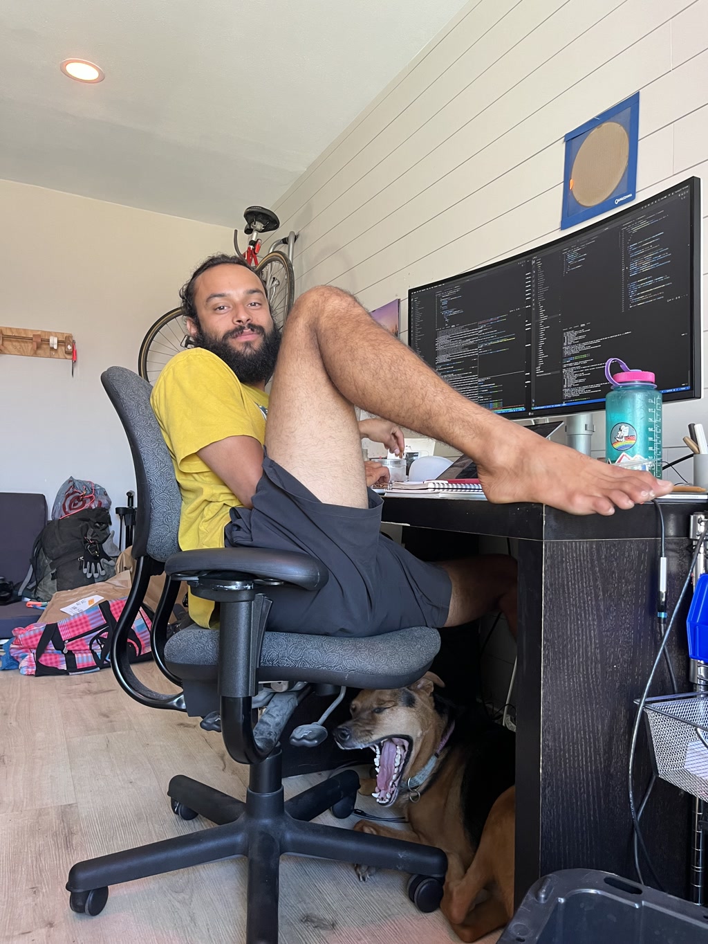 A person, presumably named CJ, is seen seated at a desk in a relaxed pose, one foot propped up on the desk, revealing bare legs. The individual has a beard and is wearing a yellow t-shirt with black shorts. In front of CJ is a large monitor displaying multiple lines of code, indicating a work or programming environment. To CJ's right is a water bottle with a cartoonish design. Underneath the desk, a dog, possibly named Moose, is lying down with its head poking out, yawning or panting, providing companionship. The room has a light wooden floor, and the walls are adorned with a bicycle hanging and a framed blue oval art piece above the desk. To the left, there's a backpack and some fabrics, suggesting a casual, lived-in workspace.