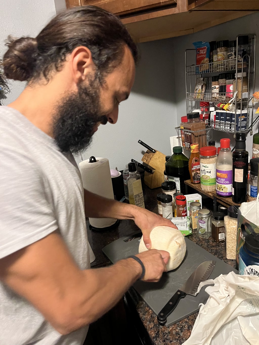 A man with a ponytail and full beard is in the process of opening a young coconut. He is using a small paring knife, applying careful pressure to the white husk of the coconut. On the black cutting board beside him lies an unused larger knife, and the countertop is crowded with an assortment of cooking ingredients and condiments, including bottles of sauces, spices, and a shelf full of various pantry items. The room indicates a kitchen setting, with visible kitchenware and appliances.