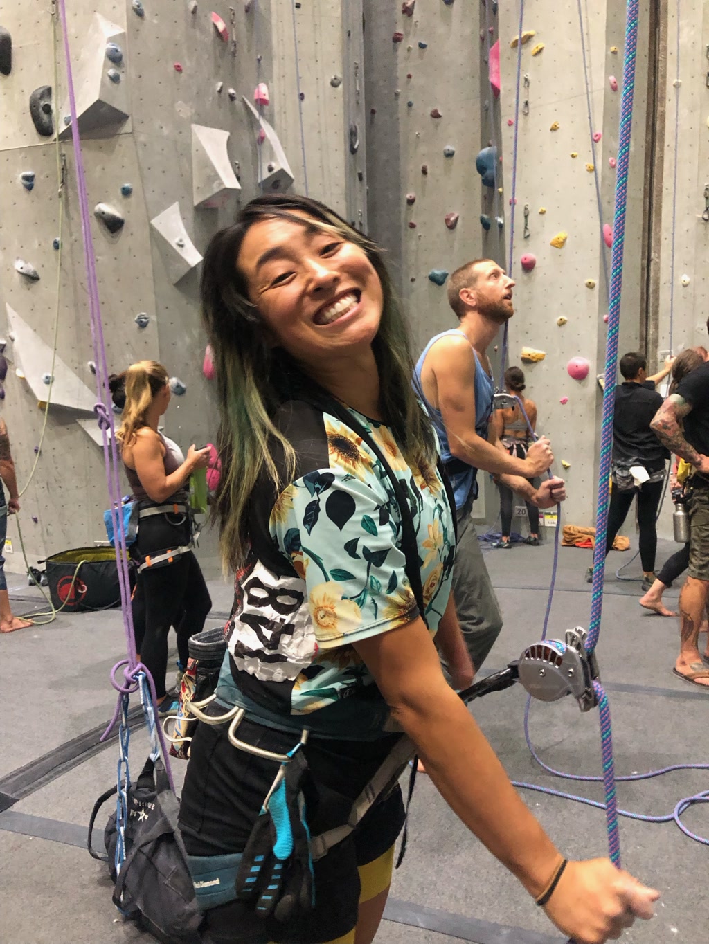 Josie is belaying at Mesa Rim, a climbing gym. She is smiling broadly and facing the camera, her energy suggesting enthusiasm and joy. She is dressed in a Hawaiian-themed shirt adorned with tropical patterns and skulls and short bottoms. Equipped with a climbing harness, she has a belay device with a rope threaded through it attached securely to her harness. Her climbing gear also includes a chalk bag attached at her back, which climbers use to keep their hands dry for better grip. Other climbers and the tall climbing walls, dotted with various colorful climbing holds, create a background bustling with indoor climbing activity.