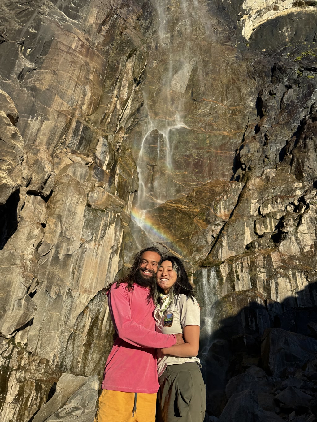 CJ and Josie are embracing warmly in front of the upper part of Bridalveil Falls in Yosemite. A bright rainbow arcs gracefully above them, casting a spectrum of colors that enhances the natural glow of their faces. They both wear happy, contented smiles. CJ has long hair and is wearing a pink long-sleeve shirt over a red t-shirt paired with yellow pants. Josie has her hair down and is dressed in an olive green t-shirt with a scouting neckerchief and khaki green pants. The sunlight highlights the misty waterfall in the background, contrasting with the rugged, dark rocky surface that forms the cliff from which the waterfall descends.