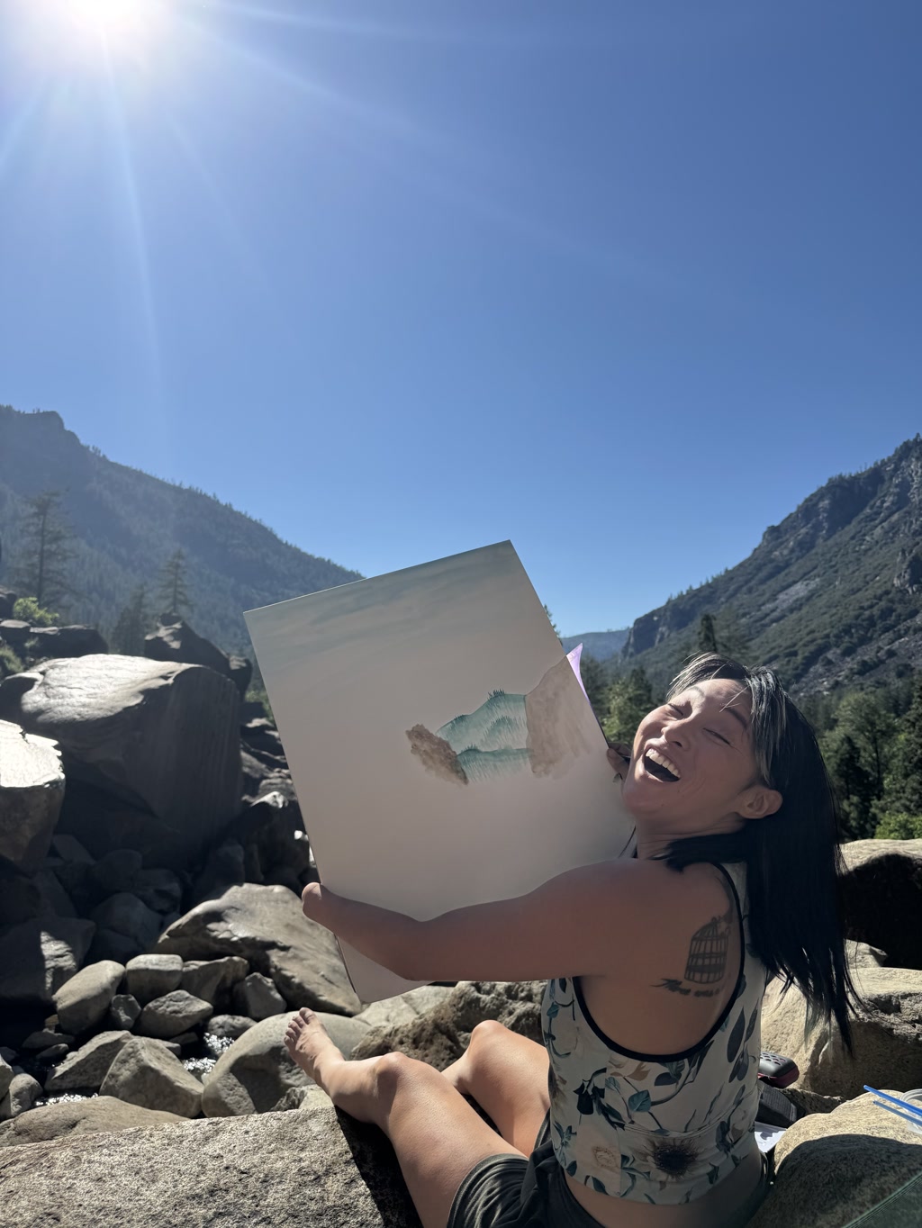 Josie sits on boulders beside a stream under a bright blue sky with the sun shining brilliantly. Lush greenery and majestic mountains are visible in the backdrop, indicative of Bridalveil falls' pools. She is holding up a piece of paper which shows the beginnings of a watercolor painting, seemingly trying to capture the surrounding landscape. Her expression is one of pure joy and excitement, and she has a tattoo on her left arm. She is wearing a sleeveless top with a botanical print and shorts, enjoying a creative moment in nature.