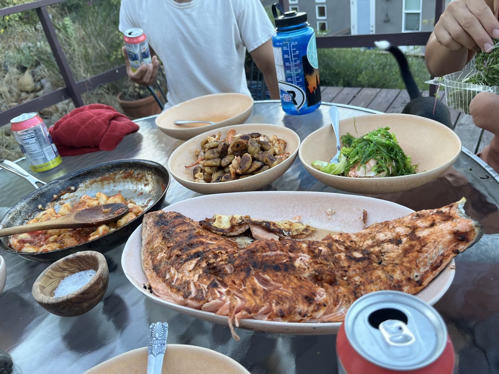 A sumptuous outdoor meal is spread out on a table consisting of a full slab of grilled salmon dominating the scene with its golden brown exterior, placed in the center on a paper-lined oval plate. To its left, there is a skillet with lobster nestled in a bed of homemade gnocchi, coated in a bright red tomato sauce. A bowl of seaweed salad with shreds of green seaweed and bits of pink, likely shrimp, occupies a paper bowl on the right, promising a refreshing taste. Accompanying these dishes are some roasted vegetables in another paper bowl, a small wooden bowl with salt, and various can drinks that suggest a relaxed dining atmosphere. There are a number of personal items like a water bottle and a red cloth, as well as two individuals partially visible, engaging in the meal, indicating a shared dining experience.