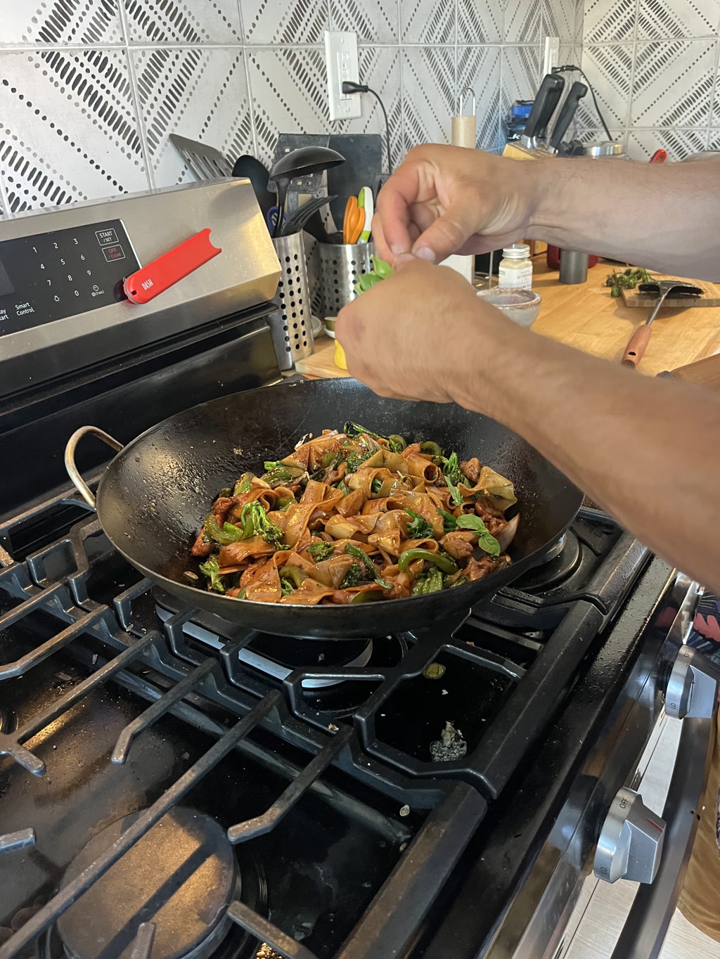 A large wok sits on a gas stove, filled with pad see ew, a Thai stir-fried noodle dish. The noodles are brown and glossy, mixed with pieces of what appears to be chicken, and green vegetables that could be broccoli. The wok emits steam, indicating that the food is hot. A person's hands are in action, tearing fresh basil leaves over the dish, adding a final touch of seasoning. The hands are well-defined and seem strong. In the background, there's a kitchen environment with various utensils and a knife block.