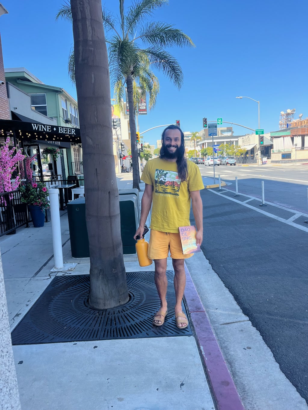 A cheerful man stands on a sidewalk, dressed in vibrant yellow and orange clothing that playfully resembles the colors of a ripe banana. He holds a bright yellow mug in one hand and a book in the other. The setting seems to be a sunny, urban area with palm trees and street signage, suggesting a warm, inviting atmosphere.